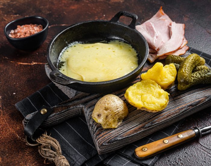 Swiss Raclette Melted cheese with boiled potato and ham on wooden board. Dark background. Top view.
